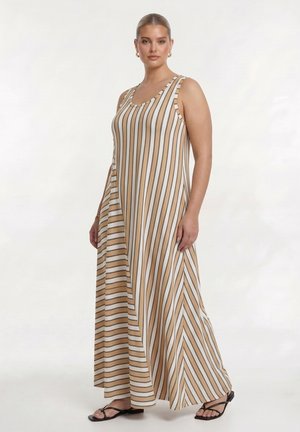Femme portant une robe longue sans manches à rayures verticales beige et blanches avec des sandales noires, debout devant un fond clair uni.