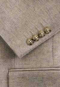 Béžový textúrovaný rukáv saka so štyrmi prišitými gombíkmi z korytnačieho vzoru a viditeľným prešitím manžety.