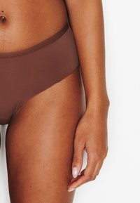 Slip bikini marrone in misto cotone con una trama liscia e una fascia elastica in vita. Vista ravvicinata che mostra l'apertura della gamba e la posizione della mano.