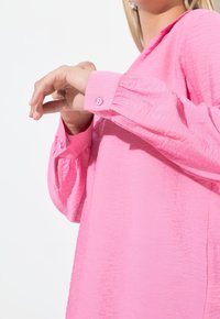 Langärmlige, pinke Bluse aus strukturiertem Stoff. Die Bündchen haben passende pinke Knöpfe, und das Hemd hat eine lockere Passform.