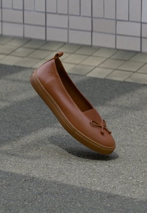 Brown leather flat shoe with a small bow detail, suspended above a pavement surface beside a tiled wall background.