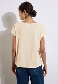 Top de algodón de manga corta y ajuste holgado en amarillo pálido. Presenta un escote amplio y mangas enrolladas, combinado con jeans azul oscuro.
