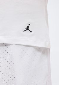 Λευκά αθλητικά ρούχα με μαύρο λογότυπο Jumpman κεντημένο κοντά στο τελείωμα του πουκαμίσου, με μέρος διάτρητων σορτς να φαίνεται από κάτω.