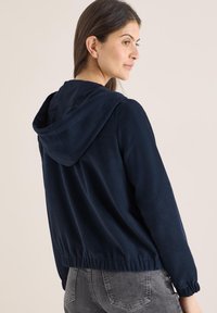 Veste polaire bleu marine avec capuche, ourlet et poignets élastiques. Texture lisse avec une coupe décontractée, vue de côté-arrière.