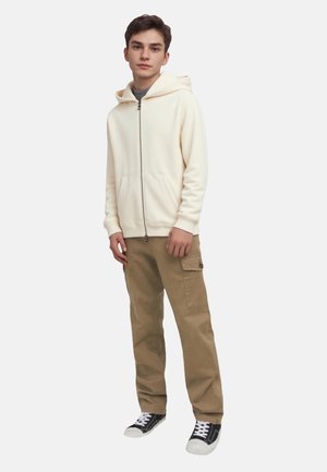 Cream zip-up hoodie with a drawstring hood, paired with beige cargo pants and black and white sneakers. Smooth fabric and relaxed fit.