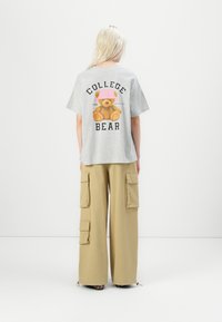 Gray cotton t-shirt with a teddy bear graphic and "COLLEGE BEAR" text on the back, paired with tan cargo pants featuring side pockets.