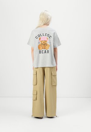 Gray cotton t-shirt with a teddy bear graphic and "COLLEGE BEAR" text on the back, paired with tan cargo pants featuring side pockets.