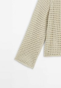 Manche en crochet beige avec motif carré ajouré et poignet uni sur fond blanc.