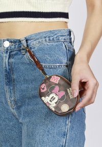 Monedero marrón con cremallera y diseño de Minnie Mouse, con acentos rosas y una correa de muñeca, sostenido contra unos pantalones vaqueros de cintura alta en azul.
