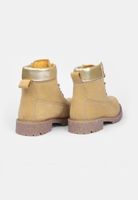 Paire de bottines en cuir marron clair avec colliers matelassés en or métallique et semelles en caoutchouc texturé, vues de derrière sur fond blanc.