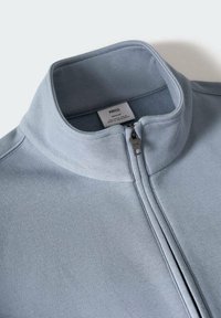 Veste zippée bleu clair avec col montant, fermeture éclair en métal visible et une petite étiquette de marque à l'intérieur du col.