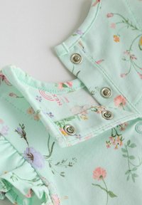 Tissu vert menthe clair avec des motifs floraux, doté de fermetures à pression et de bords froncés sur le col et les épaules. Matière en coton doux.