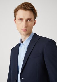 Ναυτικό σακάκι με ραφές, μονός κουμπί και μπροστινής τσέπης πάνω από ένα ανοιχτό μπλε πουκάμισο. Ομαλός ύφασμα και εφαρμογή κομμένου.