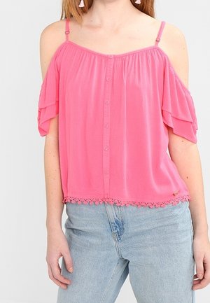 Femme portant un chemisier rose à épaules dénudées avec des boutons et un bord en dentelle, associé à un jean taille haute bleu clair.