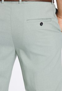 Tuvplāns uz gaiši pelēku biksēm ar melnu pogu aizmugurējā kabatā un redzamu brūnu jostu jostasvietā.