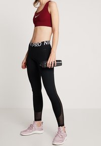 Röd sport-bh med Nike-logga, svarta leggings med nätinslag och rosa träningsskor. Håller en svart vattenflaska.