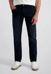 Mann in dunkelblauen Jeans und weißen Sneakern, steht mit einer Hand in der Tasche vor einem einfarbigen Hintergrund.