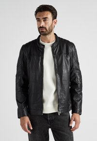 Mann, der eine schwarze Lederjacke über einem weißen Shirt trägt, kombiniert mit dunklen Jeans, steht vor einem schlichten hellen Hintergrund.