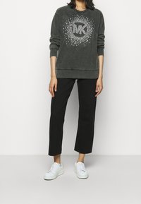 Sudadera gris oscuro con cuello redondo, que presenta un patrón de estrellas blancas y el logo 'MK' en el centro. Combinada con pantalones anchos negros y zapatillas blancas.