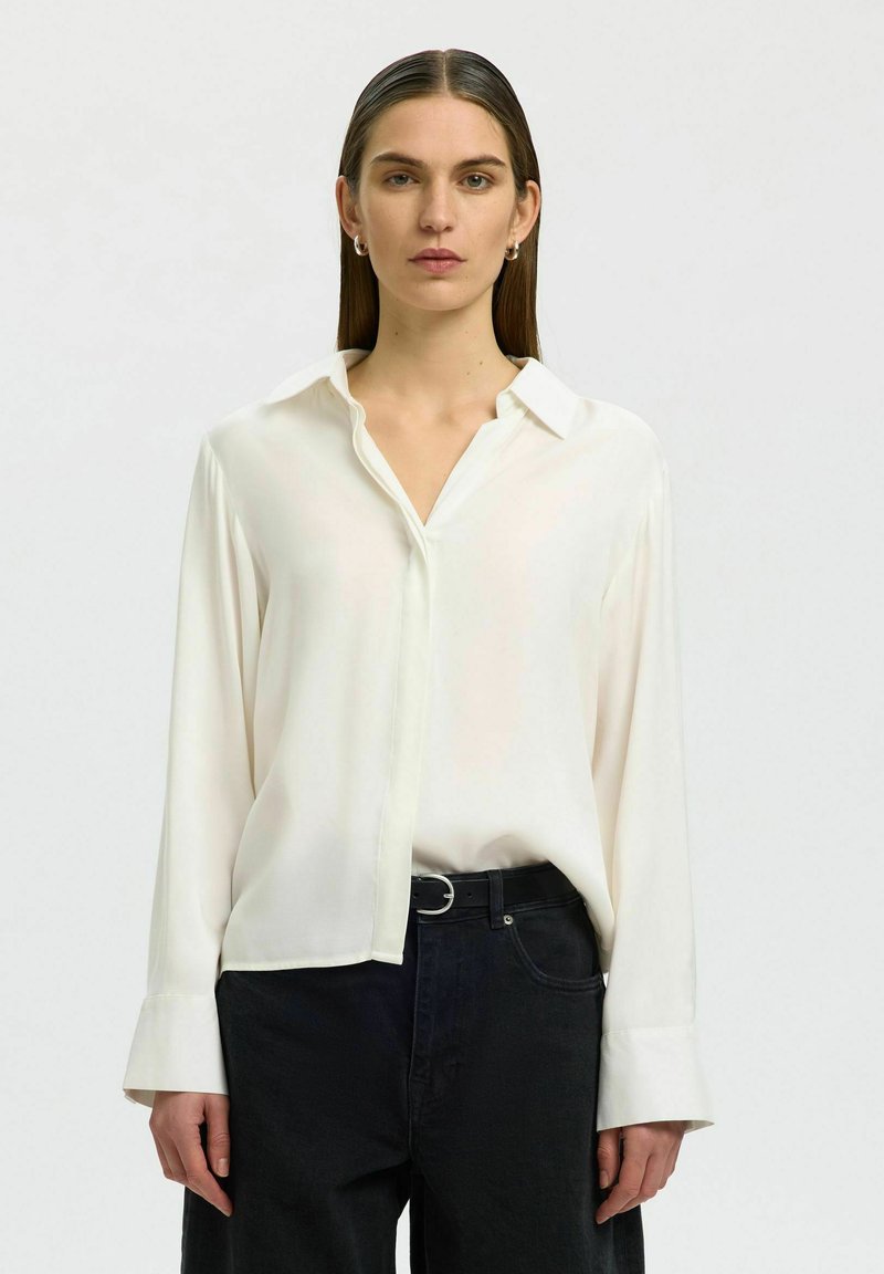 Femme aux cheveux bruns raides portant un chemisier blanc ample rentré dans un pantalon noir taille haute avec une ceinture noire, debout devant un fond uni.
