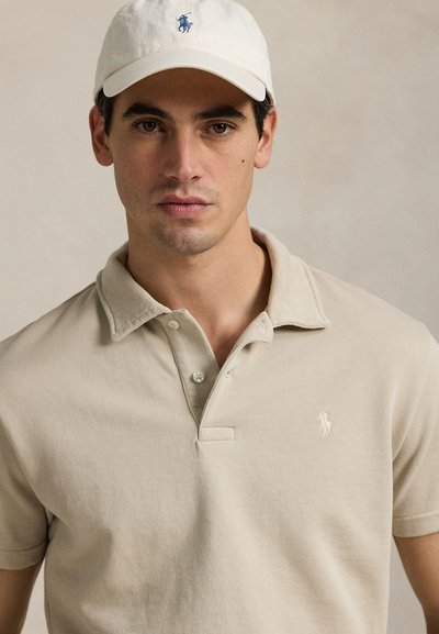 Mand iført en beige polo med logo og en matchende beige kasket, der ser lige frem mod en ensfarvet baggrund.