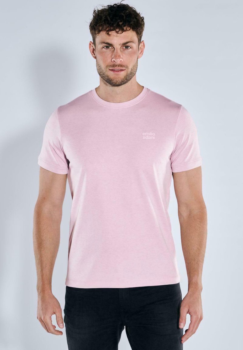 Rosa Baumwoll-T-Shirt mit Rundhalsausschnitt und kurzen Ärmeln, versehen mit einem kleinen weißen Logo auf der Brust, kombiniert mit dunklen Jeans.