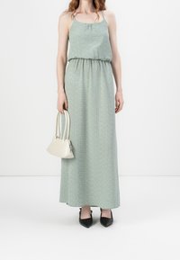 Robe longue verte à pois blancs, cintrée à la taille, avec de fines bretelles. Associée à un sac à main crème et des chaussures noires à bouts pointus.