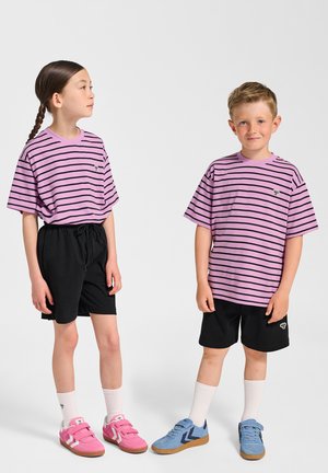 Två barn står sida vid sida. Båda bär för stora t-shirts med lila och svarta ränder, svarta shorts och sneakers i kontrasterande färger.