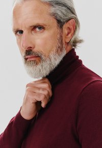 Bordowy sweter z golfem o prążkowanej fakturze, ciasno przylegający do szyi. Na zdjęciu mężczyzna poprawia kołnierzyk prawą ręką.