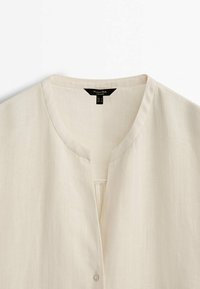 Blouse légère couleur crème avec encolure ronde et détail subtil de pli avant, fermeture à boutons partiellement visible en bas.
