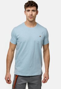 Helle blaue T-Shirt aus weichem Material, mit Rundhalsausschnitt, kurzen Ärmeln und einer Brusttasche auf der linken Seite, die mit einem kleinen runden Knopf verziert ist.