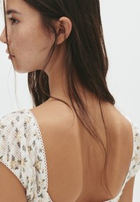 Weißes Kleid mit floralem Muster und gerafftem Design, mit einem tiefen Rückenausschnitt und zarter Spitzenverzierung an den Ärmeln. Die Haare fallen sanft über die Schulter.