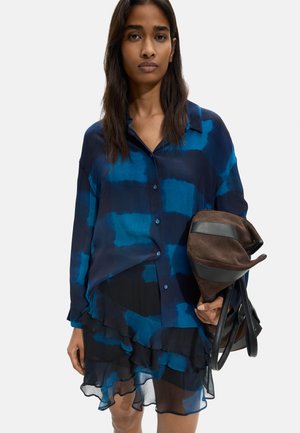 Kobieta ubrana w niebiesko-czarną koszulkę tie-dye i falbaną spódnicę, trzymająca brązową i czarną torbę w lewej ręce.