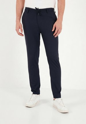 Homme portant un pantalon slim bleu marine avec cordon et des baskets blanches, debout devant un fond clair uni.
