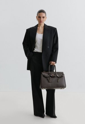 Woman in black oversized blazer and wide pants holds large dark brown handbag, standing against plain light gray background.