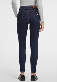 Jeans ajustados de color azul oscuro con una cintura alta, que cuentan con bolsillos traseros y un parche de logo de cuero en la cinturilla. Textura de denim suave.