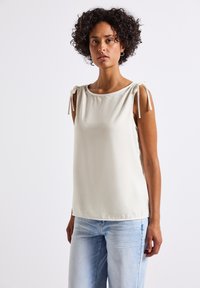 A woman with curly hair wears a light-colored sleeveless top with shoulder ties and light blue jeans, standing against a plain background.