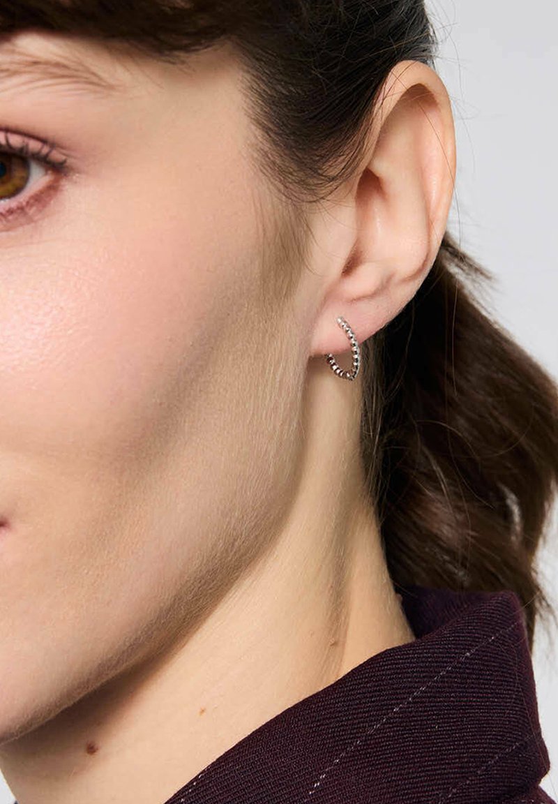 Srebrna kolczyk w kształcie obręczy z teksturowaną powierzchnią, z spiralnym wzorem. Noszony na płatku ucha, z widocznymi naturalnymi włosami i skórą.