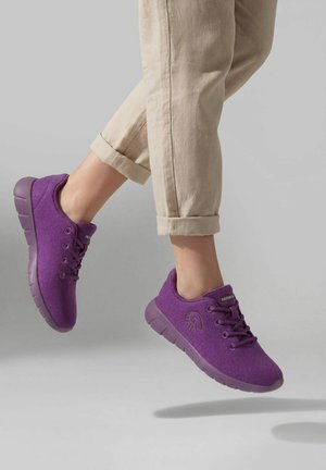 Pieds portant des baskets violettes à lacets et un pantalon beige roulé, en plein saut sur un fond gris clair uni.