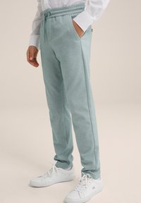 Světle modro-zelené tepláky z jemné texturované látky s elastickým pasem a bočními kapsami, doplněné bílými teniskami.