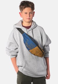 Marsupio con sezioni blu e marrone, con "baagah" ricamato in oro. Il modello indossa un hoodie grigio e pantaloni cargo scuri. Cintura regolabile.