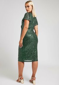 Vihreä paljettimekko, jossa on lyhyet lepattelevat hihat ja vartaloa myötäilevä siluetti, ulottuen puoleen vasikkaan, yhdistettynä kultaisiin korkokenkiin.