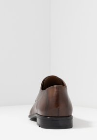 Scarpa da uomo in pelle marrone con una finitura liscia, punta arrotondata e suola in gomma nera. Presenta sottili dettagli di cucitura lungo il tallone.