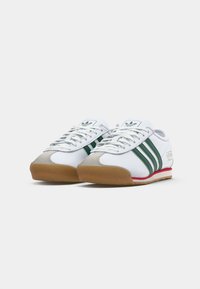 Zapatillas blancas con rayas verdes, acentos de ante y suela de goma. Presentan un cierre de cordones en la parte delantera y una marca en el lateral. Diseño plano.