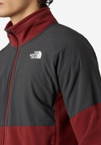 Hombre vistiendo una chaqueta de forro polar de North Face en color burdeos y gris oscuro, con cremallera frontal, logo visible en el pecho y cuello alto.