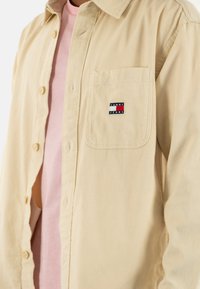 Camicia in corduroy beige con chiusura a bottoni, taschino singolo e piccolo logo multicolore "TOMMY JEANS" sul petto. Indossata sopra una maglietta rosa.