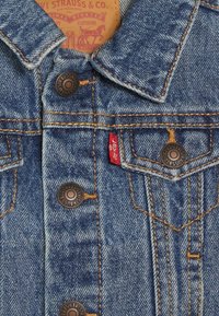 Chaleco de mezclilla de color azul claro, con botones de cobre, costuras naranjas y una etiqueta roja de Levi's. Presenta un cuello puntiagudo y dos bolsillos en el pecho.
