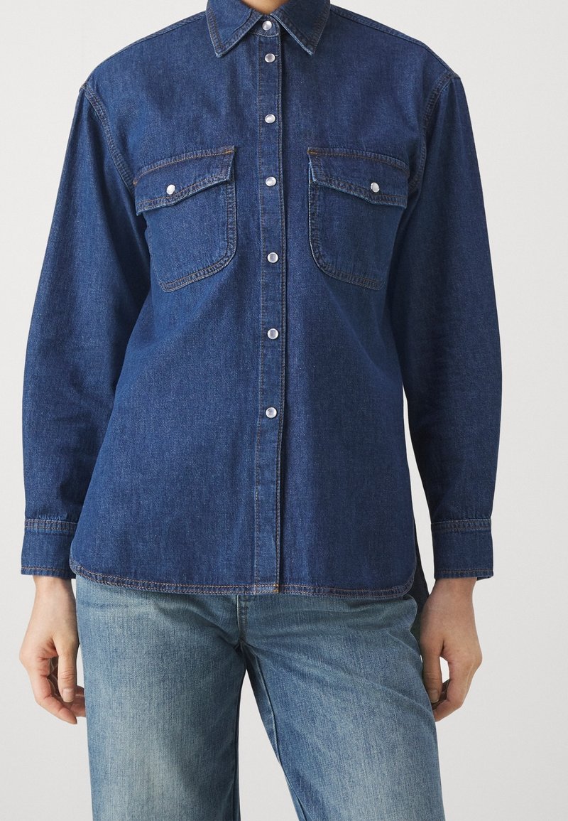 Mujer con camisa vaquera azul oscuro abotonada con dos bolsillos en el pecho combinada con vaqueros vaqueros azul claro.