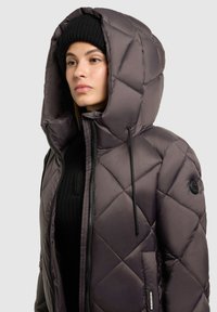 Gequiltte donkergrijze jas met een grote capuchon, een voorzak en zwarte trekkoorden. Draagt een geribde zwarte trui eronder.