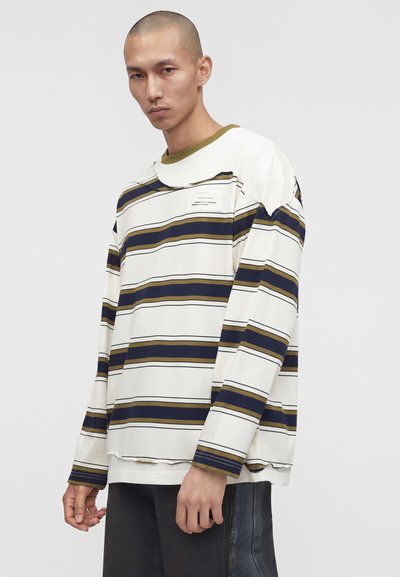 Άνδρας φοράει φαρδιά λευκή μακριά μπλούζα με οριζόντιες ναυτικές και ελαιώδεις ρίγες, με ελαφρώς φθαρμένο γιακά και τελείωμα, στέκεται μπροστά σε απλό φόντο.